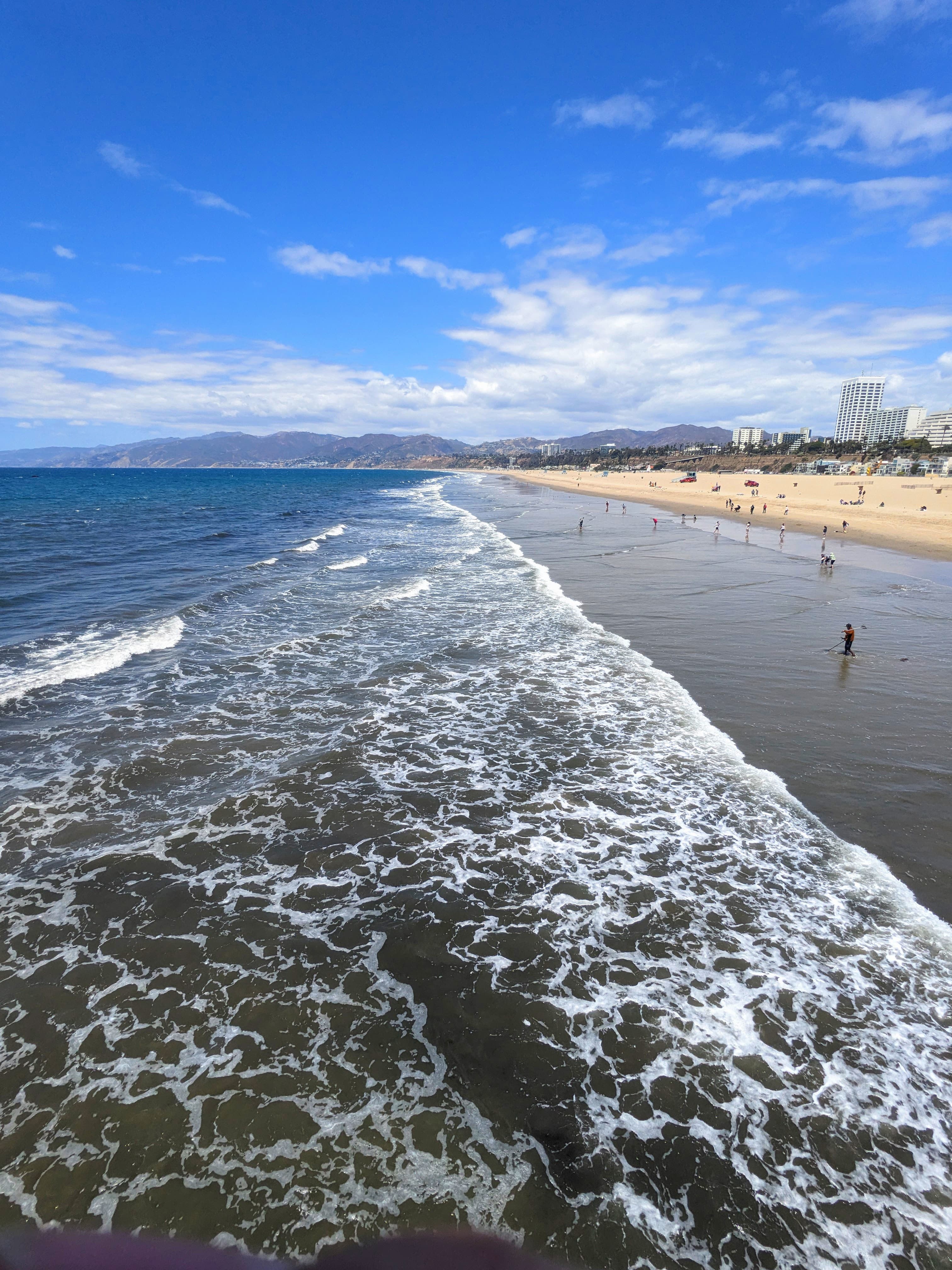 Foamy waves roll onto a sandy beach with distant mountains and buildings under blue skies.