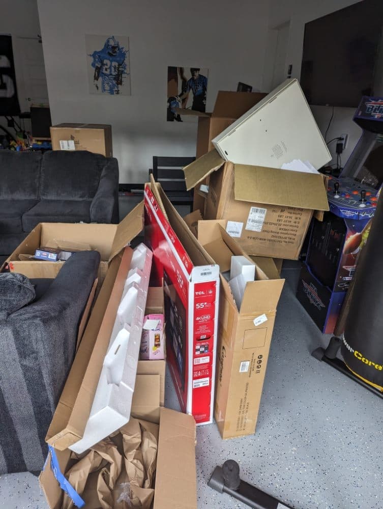 Cluttered garage filled with cardboard boxes, a red TV box, grey sofa, and arcade machine.