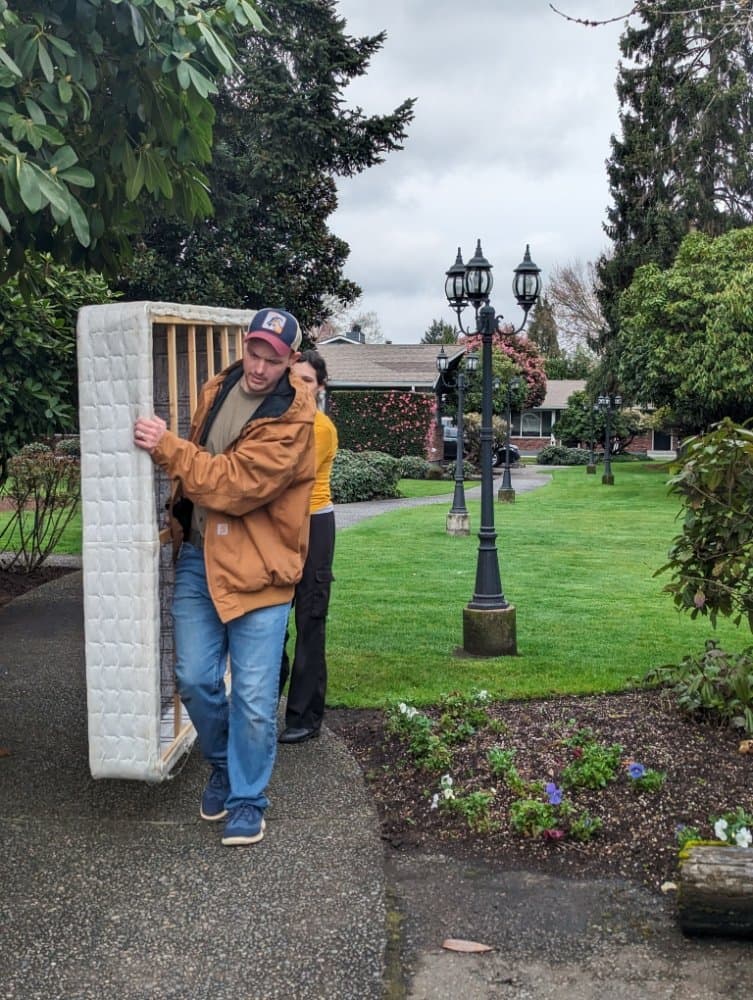 A man in a brown jacket carries a mattress along a paved residential walkway.