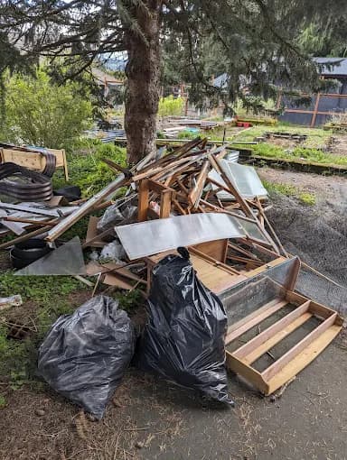 Scrap wood, glass panels, and black trash bags piled outdoors beneath a large pine tree.