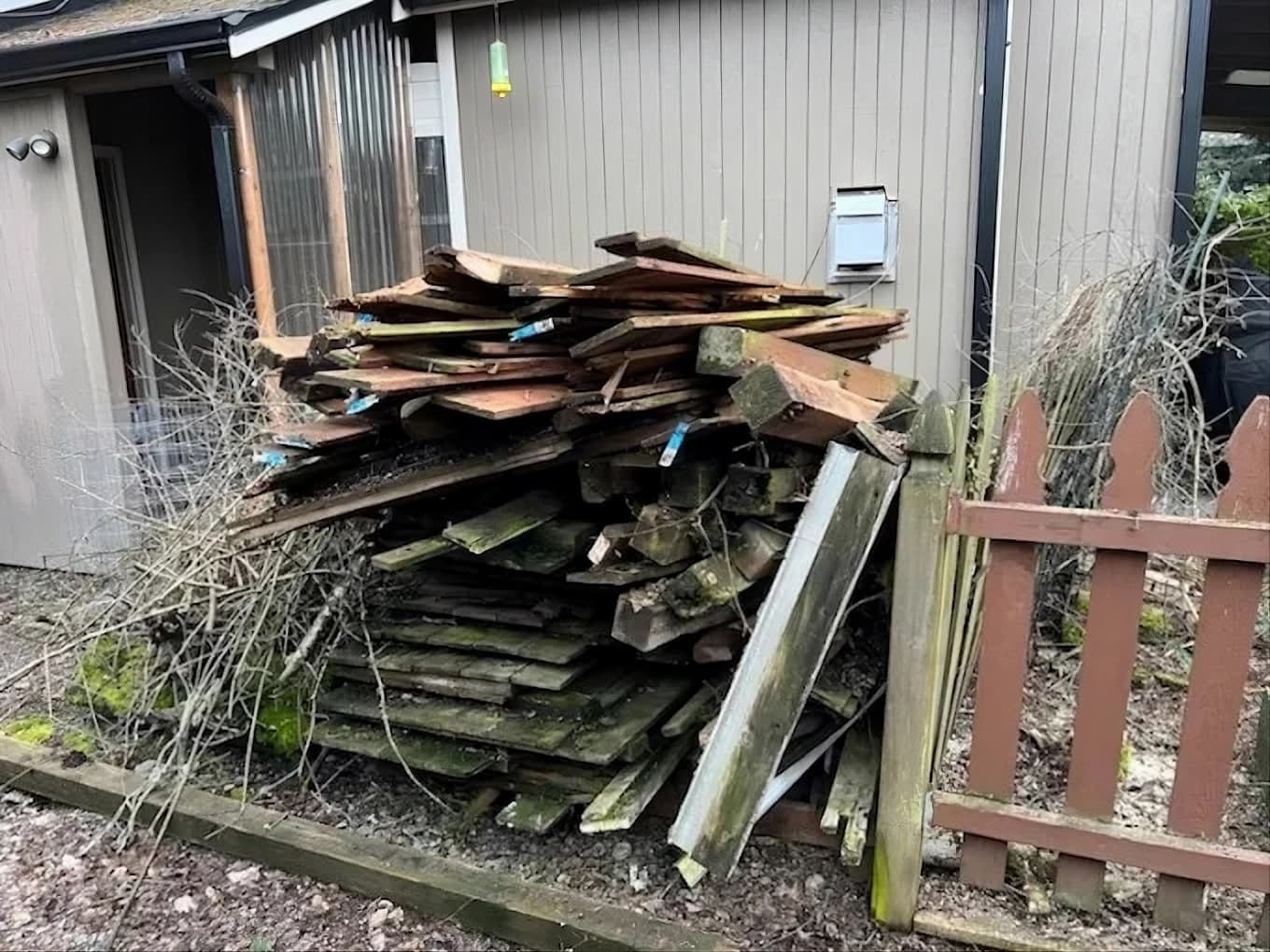 Weathered lumber and scrap wood piled high against a building beside a brown picket fence.